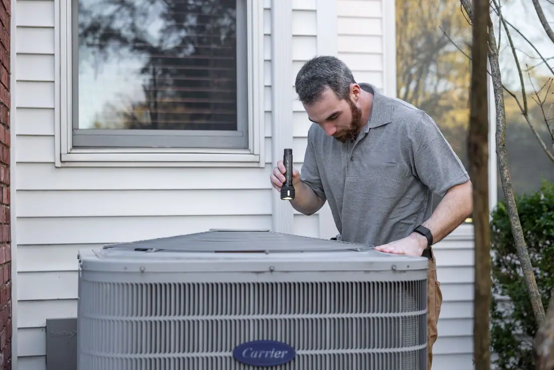 A technician inspecting an outdoor central air condenser beside a sided house